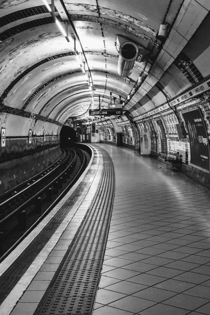Black and white image of an underground station platform with curved tiled flooring, metal rails for the train tracks, and a tiled ceiling with exposed pipes and ventilation ducts. The platform appears clean with no visible dust or debris, and the lighting evenly illuminates the area, highlighting the smooth, shiny surface of the tiles. The station walls have advertisements and directional signs, and there are no people present. This setting showcases a well-maintained environment suitable for regular surface cleaning and sanitation by companies like elephantandcastlecarpetcleaning.co.uk, emphasizing the importance of hygiene and upkeep in public transit spaces.