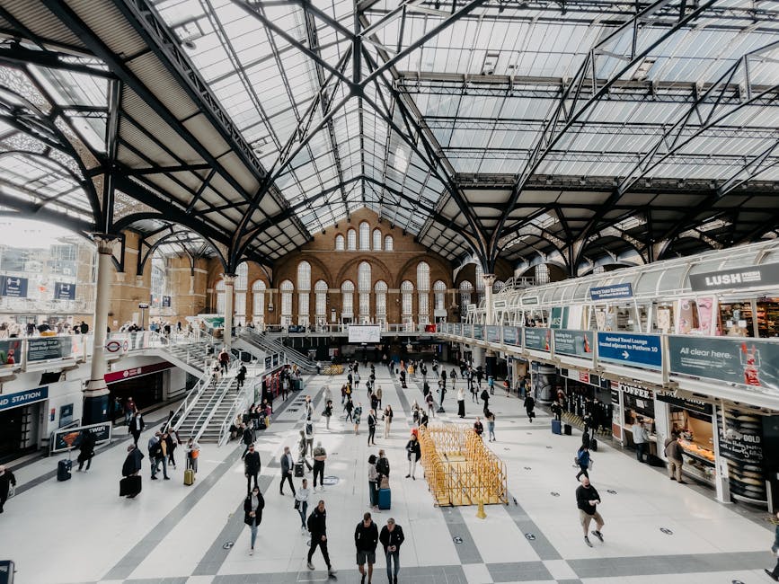 A wide view of Elephant and Castle station showcasing its historic architectural façade with ornate stonework and decorative elements. The station entrance features an archway through which pedestrians are walking across a paved, clean, and open public square. To the left, there is a modern glass structure, and on the right, a small kiosk with bright pink and yellow signage. The scene is illuminated by natural daylight, with clear blue sky above, emphasizing the tidy and well-maintained environment reflecting routine surface cleaning and upkeep. The image promotes the importance of hygiene and maintenance in public spaces, in line with services provided by elephantandcastlecarpetcleaning.co.uk.