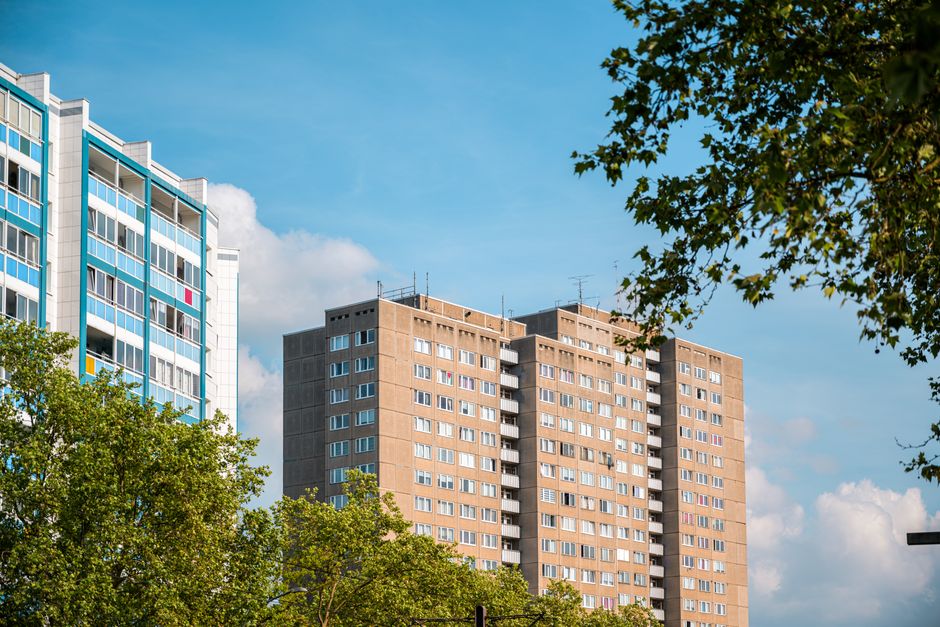 Photograph of high-rise residential buildings with modern windows, surrounded by green trees under a bright blue sky with some clouds. One building features a blue and white facade with balconies, while another has a brown brick exterior with numerous windows. The scene is taken during daytime with natural lighting highlighting the clean surfaces of the buildings and lush foliage. The photograph represents urban living with a focus on building architecture and exterior environment, consistent with the surface cleaning and maintenance themes of elephantandcastlecarpetcleaning.co.uk's Heygate Estate upholstery cleaning page.