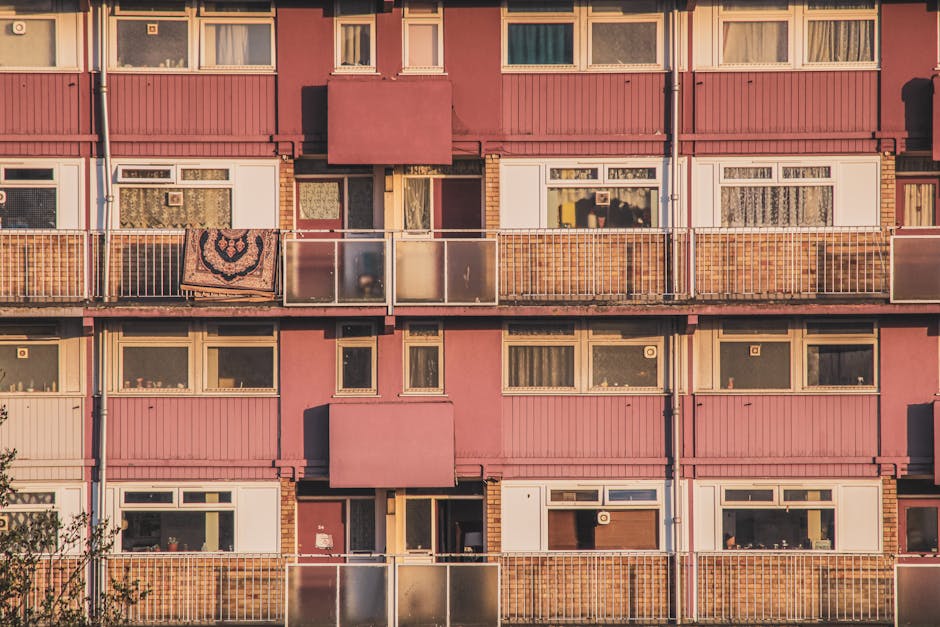 Close-up view of the exterior of a pink multi-story residential building on the Heygate Estate in Elephant and Castle, featuring numerous windows, some with curtains and blinds, and small balconies with metal railings. One balcony has a decorative blanket draped over the railing, and the building's facade showcases a mix of brick and paneling, illuminated by natural light. This image, associated with elephantandcastlecarpetcleaning.co.uk, emphasizes the urban residential setting related to cleaning and maintenance services on the estate.