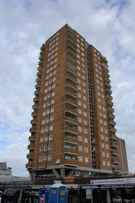 Photograph of high-rise residential buildings with modern windows, surrounded by green trees under a bright blue sky with some clouds. One building features a blue and white facade with balconies, while another has a brown brick exterior with numerous windows. The scene is taken during daytime with natural lighting highlighting the clean surfaces of the buildings and lush foliage. The photograph represents urban living with a focus on building architecture and exterior environment, consistent with the surface cleaning and maintenance themes of elephantandcastlecarpetcleaning.co.uk's Heygate Estate upholstery cleaning page.